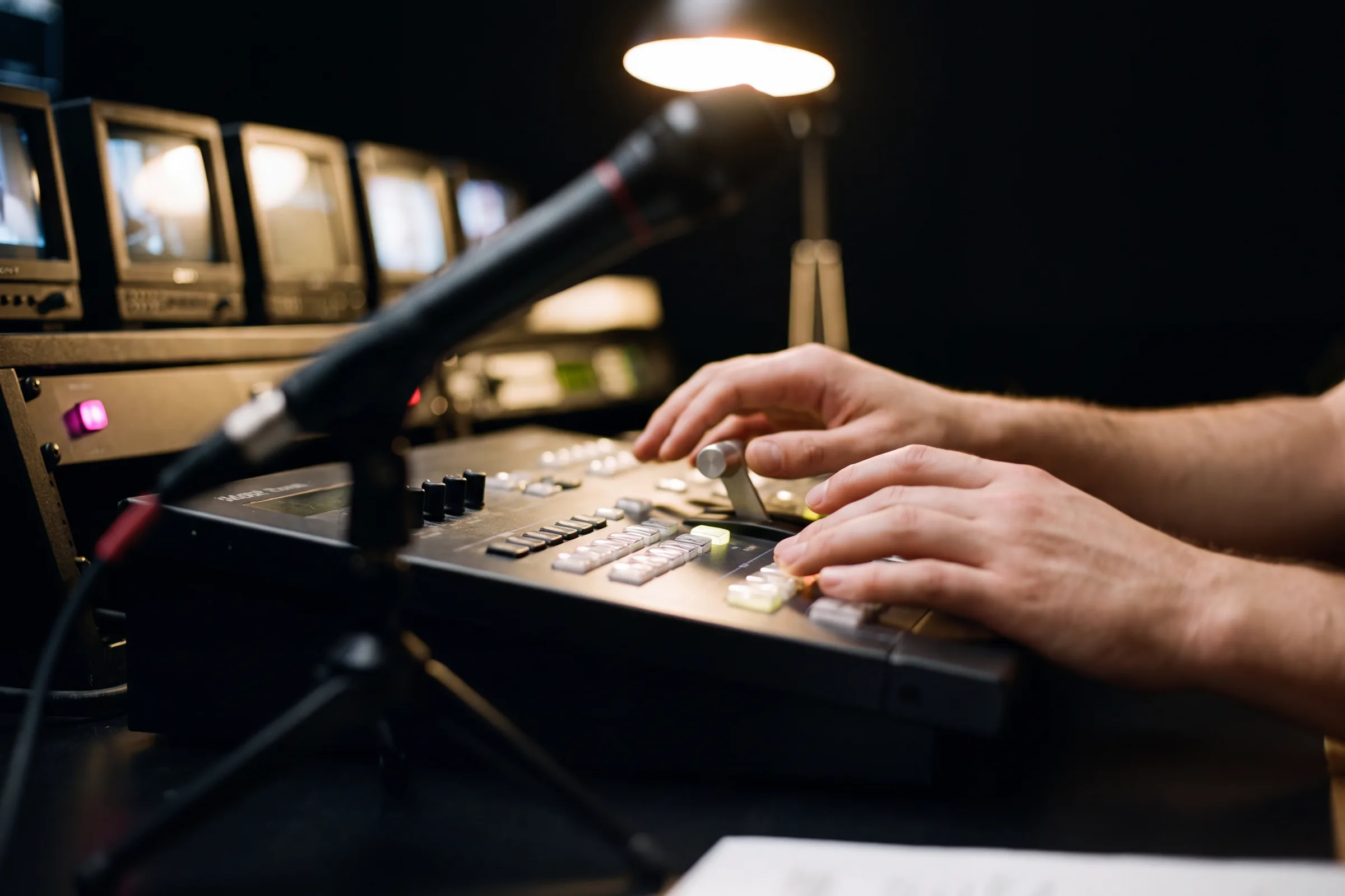 Karanlık yayın odasında switcher üzerindeki eller, arkada odak dışı önizleme monitörleri